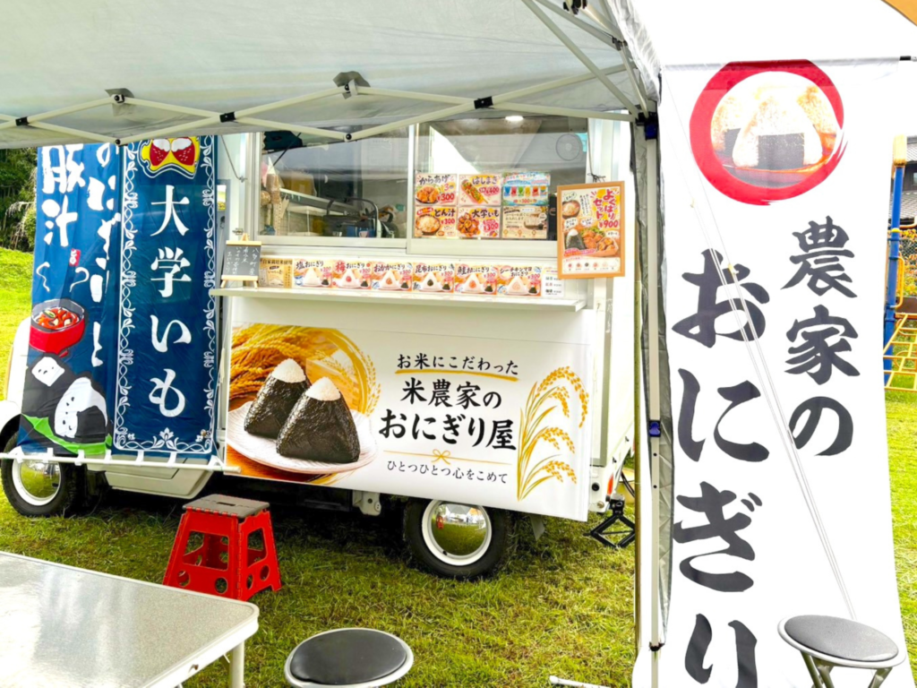 屋号：etudes（エチュード）のキッチンカー営業中の画像です。運営母体はK.Farmer（ケーファーマー）です。ケーファーマーが育てた米で作るおにぎりを販売しています。その他メニューの豚汁や大学いもなど、メニュー表やのぼりが並んでいます。